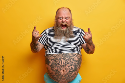 Indoor shot of overweight rock fan gestures actively and shouts, enjoys favorite music, keeps mouth widely opened, shows horns hand gesture, has very big tattooed belly, stands against yellow wall
