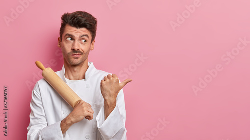 Obraz na płótnie Serious inexperienced chef points thumb aside on copy space, shows information for your recipe or menu, dressed in uniform, holds wooden rolling pin, pink background