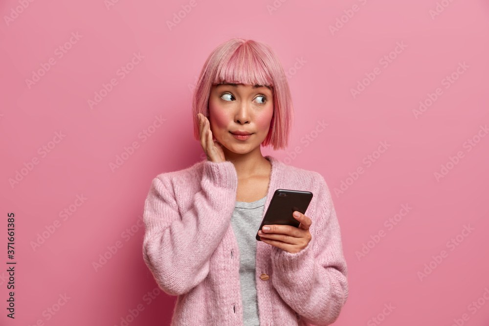 Young Asian woman with surprised face expression looks aside, dressed in casual wear, holds cellphone, surfes social media, sends content shares multimedia online, isolated over rosy background Young Asian woman with surprised face expression looks aside, dressed in casual wear, holds cellphone, surfes social media, sends content shares multimedia online, isolated over rosy background