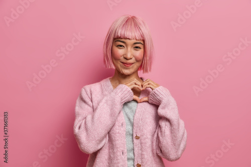 Kind positive amiable Asian woman with bob hairstyle, makes heart shape gesture, shows love care to boyfriend, generosity symbol, wears casual pink jumper poses indoor. Devotion and affection concept.