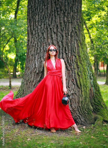 Attractive young girl, lady in long red dress in summer forest park, fashionable woman outdoors in nature