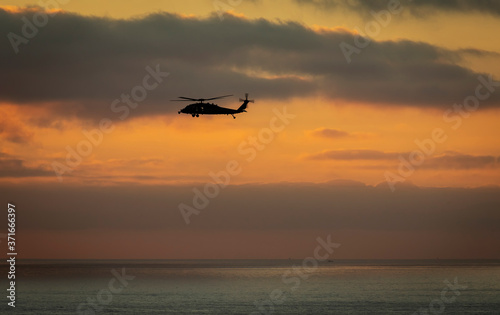 A military helicopter flying toward the left with a cloudy sunset over the pacific ocean