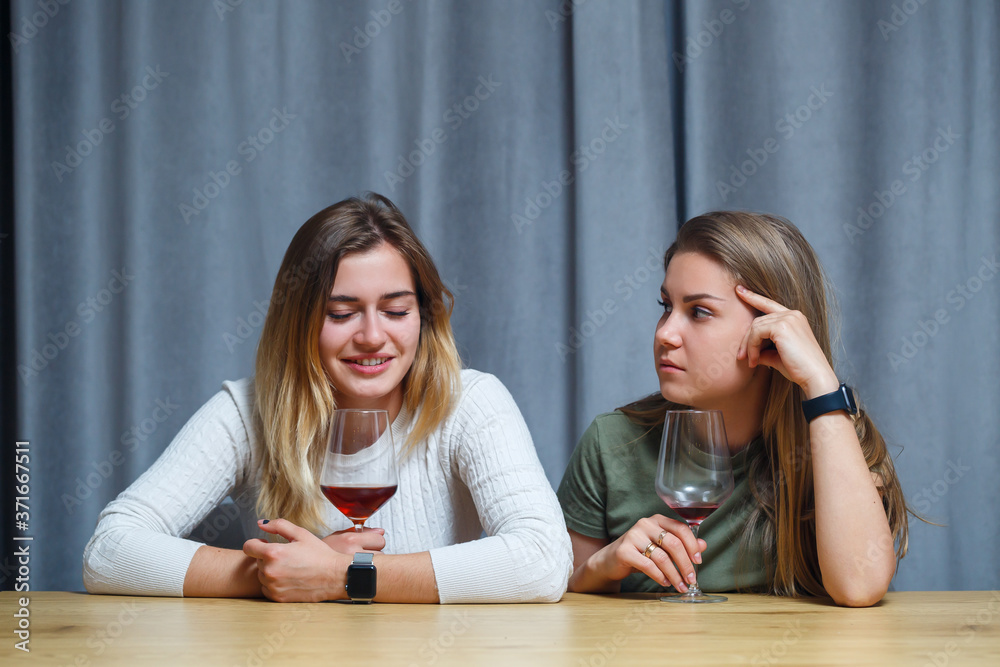 Two best friends. Young European-looking women having a conversation sitting at a desk holding a glass of wine. beautiful girls sisters love to drink alcohol laughing cheerful relax in the living room Two best friends. Young European-looking women having a conversation sitting at a desk holding a glass of wine. beautiful girls sisters love to drink alcohol laughing cheerful relax in the living room