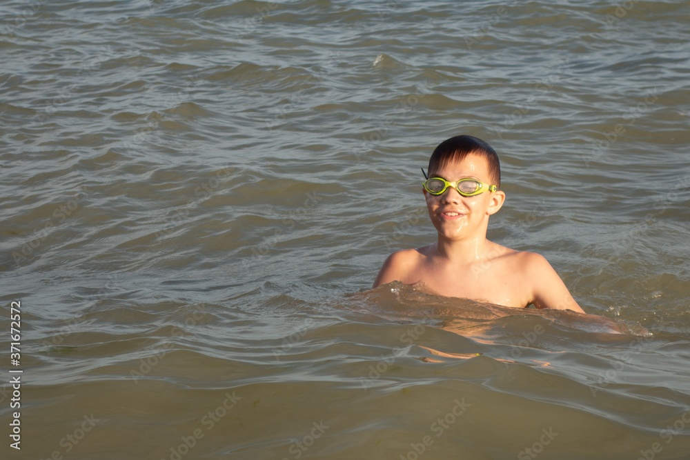 A 10-year-old boy is swimming and having fun in the sea near the shore in green swimming goggles.