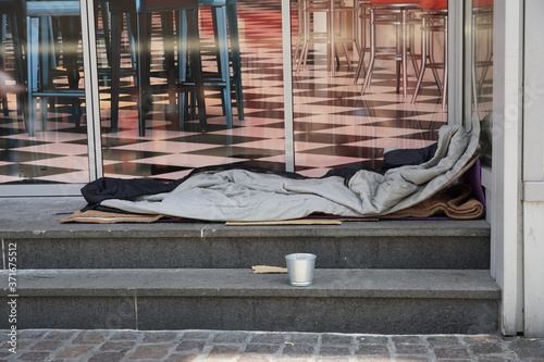 Fototapeta Naklejka Na Ścianę i Meble -  Sleeping bag of a homeless person spread on the stairs in front of a modern posh lounge or restaurant. There is a small metal cup near the berth asking for alms and charity.