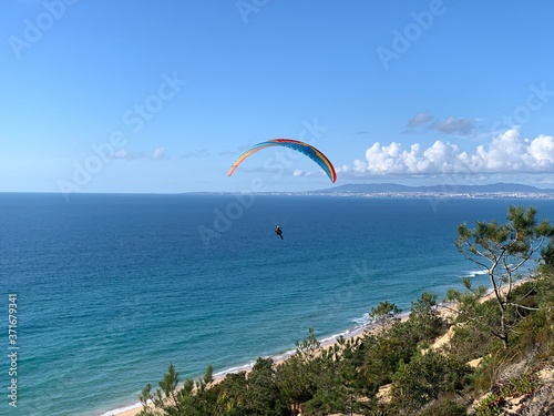 Beautiful colorful paragliding sail above a landscape with green pine tree, sand and green dunes vegetation under a beautiful blue sky in Fonte da Telha, Costa da Caparica, Portugal