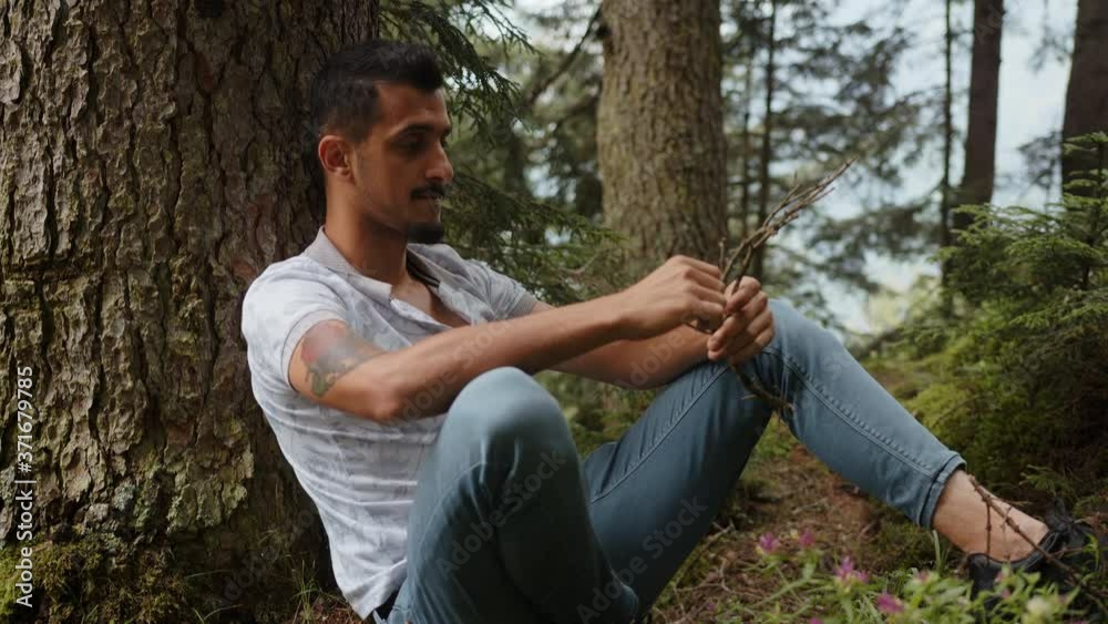Handsome young man leaning on tree is dealing with wood stick in his hand, relaxation concept Handsome young man leaning on tree is dealing with wood stick in his hand, relaxation concept