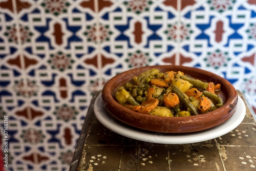 Moroccan vegetable tajine on wooden table, with traditional arabic tiles in the background.