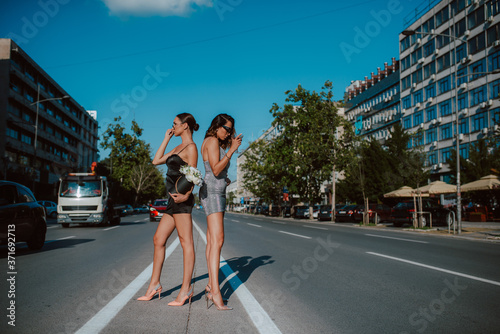 Two attractive caucasian women in summer tight dresses pose with flower boxes and cocktail on the road. Fashion and the city