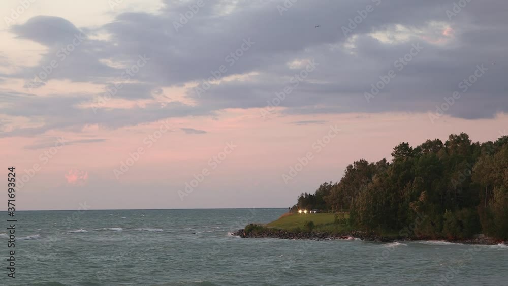 Scenic coastal island at dusk with waves crashing, cars driving along the road with headlights and seagulls in the air. Wide static. Lake Huron. Scenic coastal island at dusk with waves crashing, cars driving along the road with headlights and seagulls in the air. Wide static. Lake Huron.