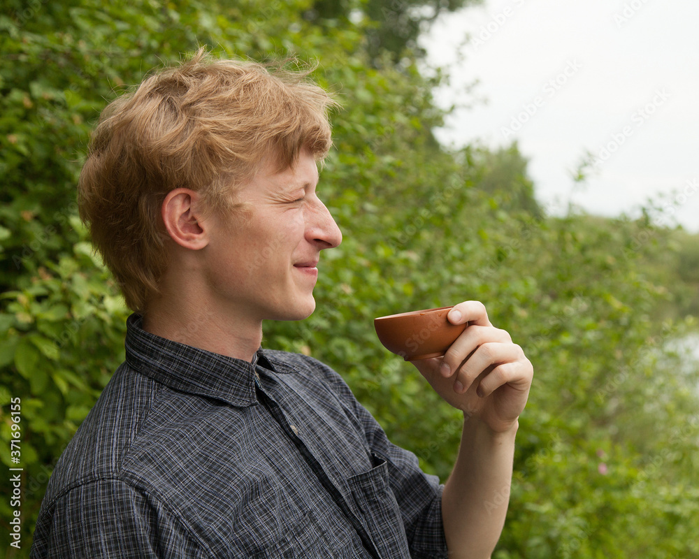 Fototapeta premium Young man with blonde hair drinking chinese tea in summer park. Closeup portrait.
