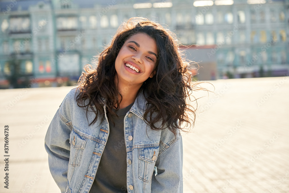 Happy African young woman wearing denim jacket laughing looking at camera standing on street. Smiling Afro American teen generation z hipster girl posing outdoor backlit with sunlight, portrait. - obrazy, fototapety, plakaty 