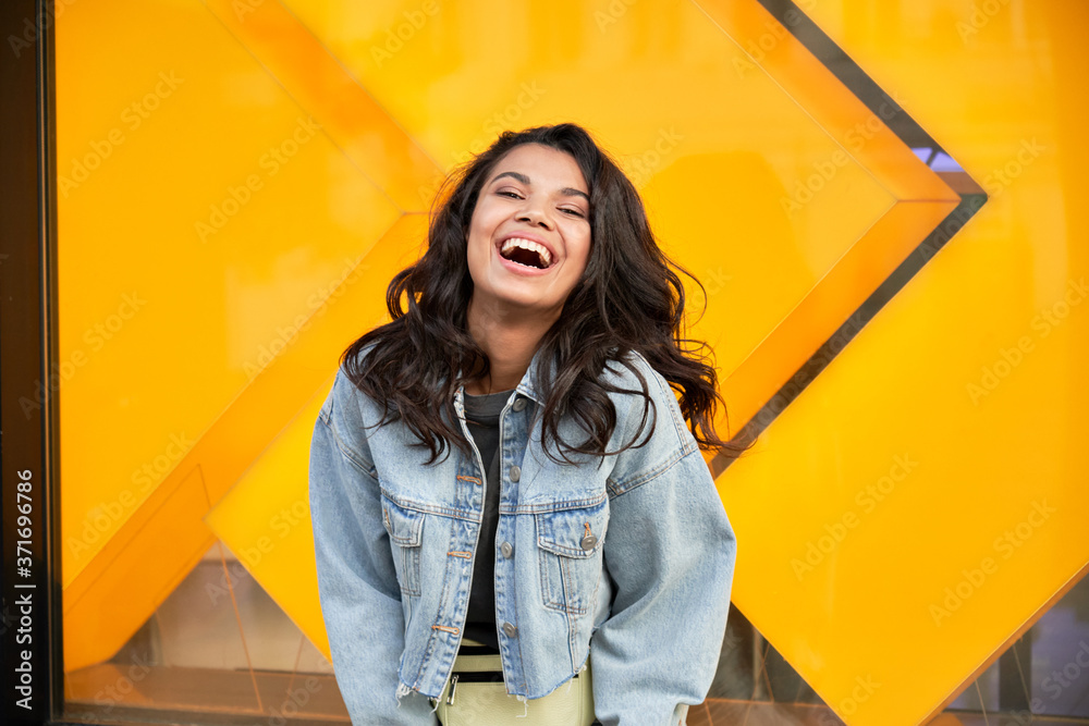 Happy African American young woman wearing denim jacket laughing looking at camera standing near city street building. Smiling positive mixed race teen generation z hipster girl posing outdoor. - obrazy, fototapety, plakaty 