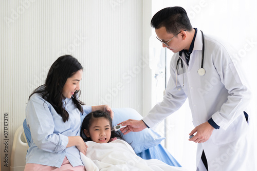 asian doctor using digital thermometer to measure temperature of young girl who is lying in the bed with mother in hospital pediatric ward. pediatrician and healthcare concept