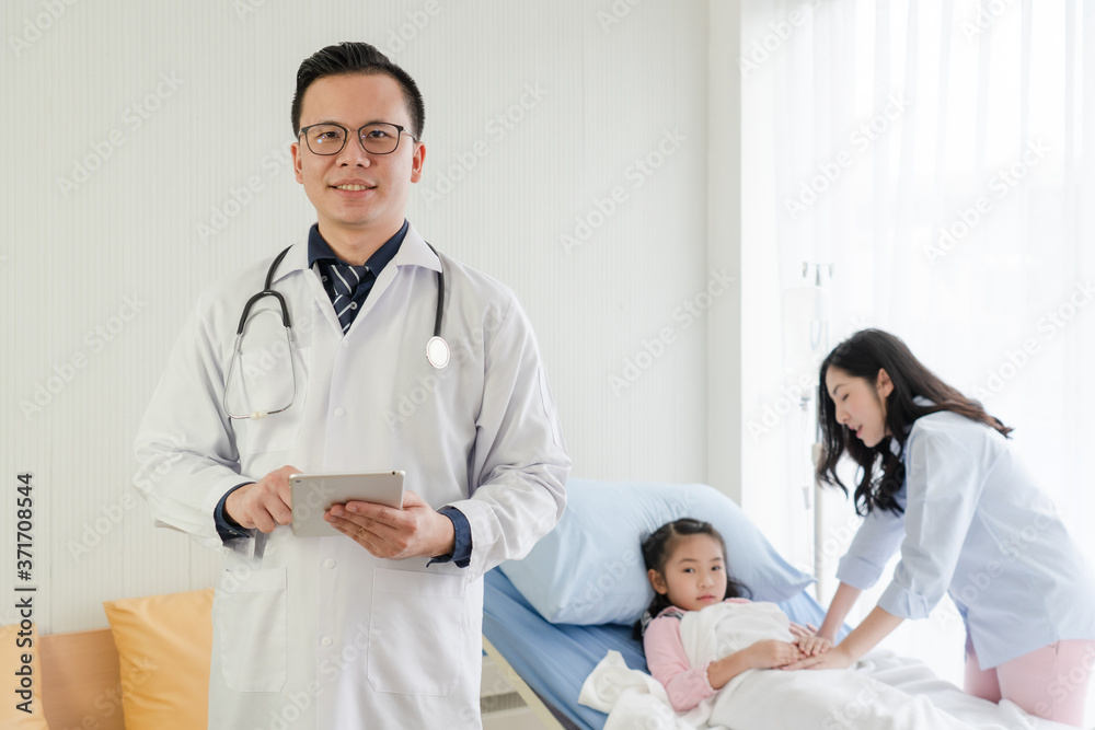 young asian doctor wearing white coat, holding tablet and standing in front of mother and girl lying on bed in hospital ward. healthcare and medical concept young asian doctor wearing white coat, holding tablet and standing in front of mother and girl lying on bed in hospital ward. healthcare and medical concept