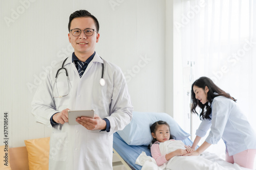 young asian doctor wearing white coat, holding tablet and standing in front of mother and girl lying on bed in hospital ward. healthcare and medical concept