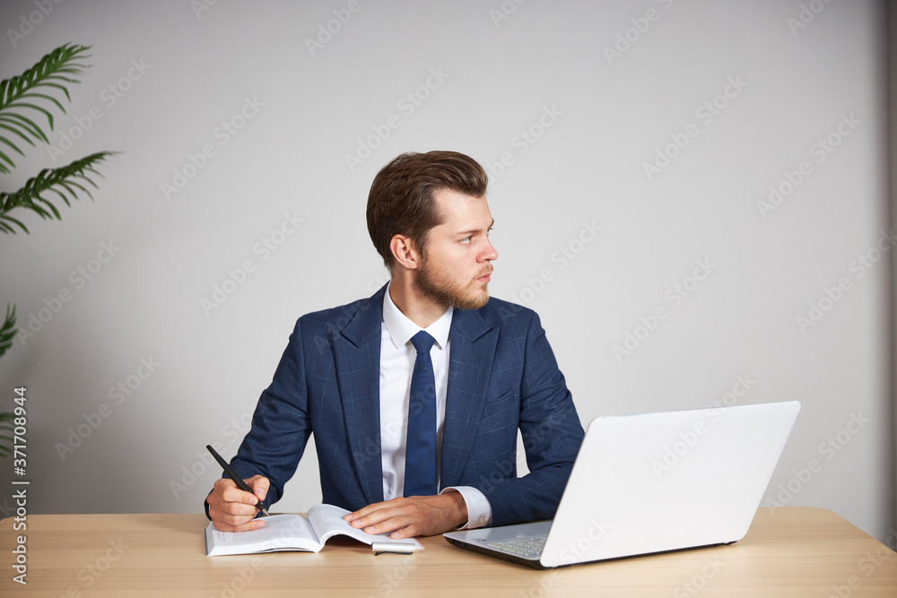 A man businessman in a blue business suit sits at a desk with a laptop, looks to the side and ponders. White background. A man businessman in a blue business suit sits at a desk with a laptop, looks to the side and ponders. White background.