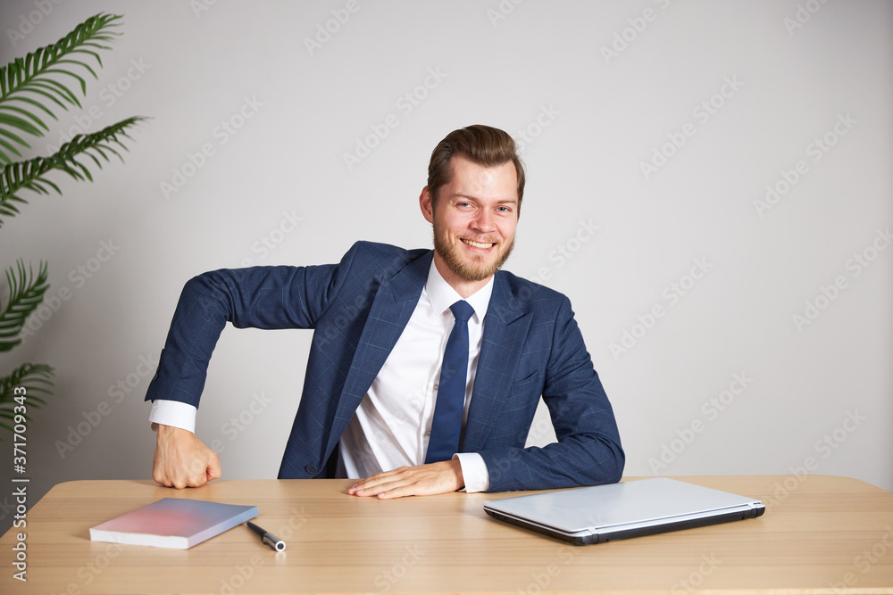 A businessman in a blue business suit sits at his desk clutching his hand in his fist. He looks directly into the camera and smiles. White background. A businessman in a blue business suit sits at his desk clutching his hand in his fist. He looks directly into the camera and smiles. White background.