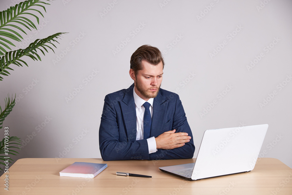 A businessman in a blue business suit sits at a wooden desk. He crossed his arms and looks into a laptop with a serious face. A businessman in a blue business suit sits at a wooden desk. He crossed his arms and looks into a laptop with a serious face.