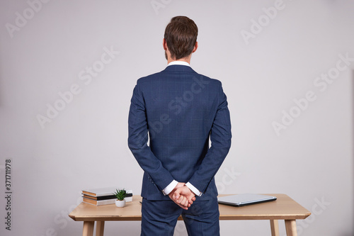 A businessman in a blue business suit stands in front of an office wooden table with his back to the camera with his fists clenched.