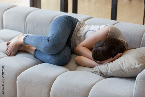 Frightened young woman lying on sofa at home curled up in fetal position, sleeping on couch in living room feeling terrible tired or stomachache, hiding from troubles, having psychological problems