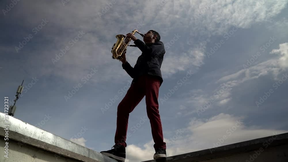 The silhouette of a young stylish guy playing a saxophone on the roof of a high-rise building against the sky. Wide view from below The silhouette of a young stylish guy playing a saxophone on the roof of a high-rise building against the sky. Wide view from below
