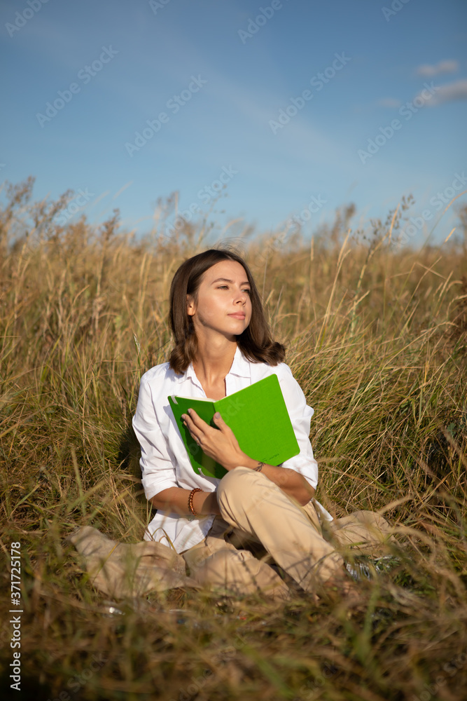 Young European student woman with a book sitting in a field in autumn or summer Young European student woman with a book sitting in a field in autumn or summer