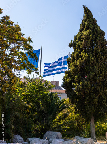 the Greek flag flies on a pole. the wind blows the canvas of the Greek flag. flag in the center of athens. flag among the trees on the background of the acropolis