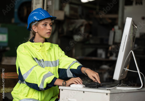 Portrait of young beautiful technician woman or industrial worker with hard hat or helmet and vest working electronic machine on notebook and computer in Factory of manufacturing place 