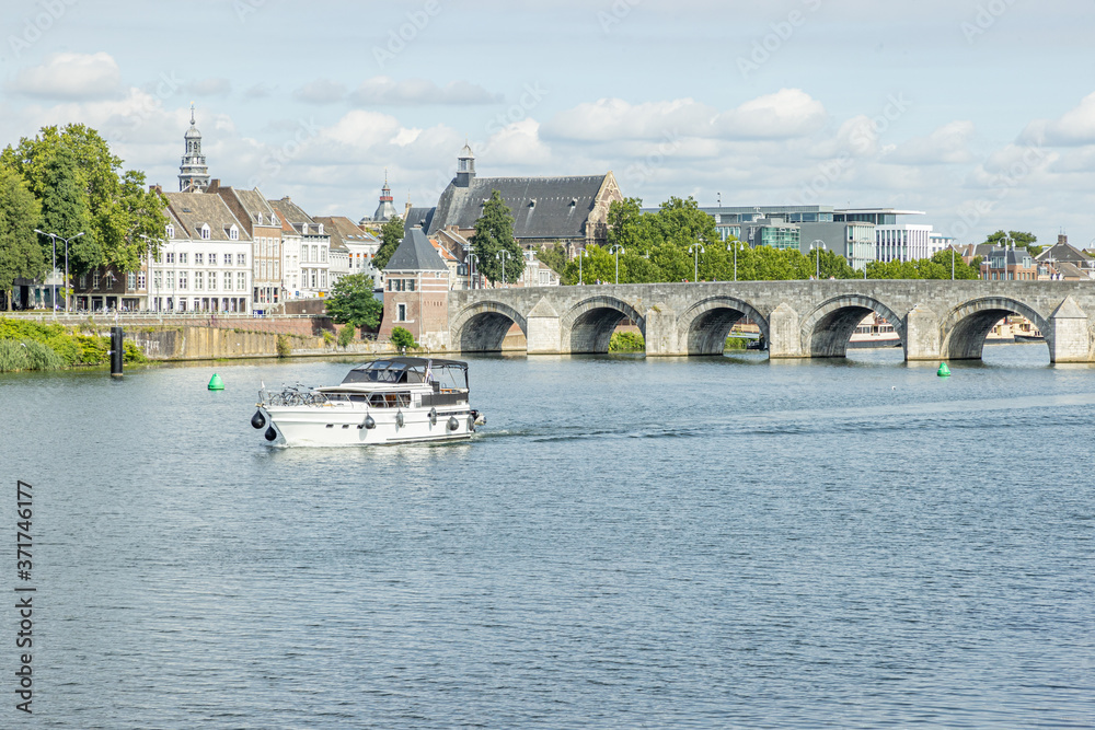 Naklejka premium Maas river with a boat sailing, the Servatius bridge (Sint Servaasbrug) and with an urban landscape of the city of Maastricht in the background, sunny day in the South Limburg, Netherlands