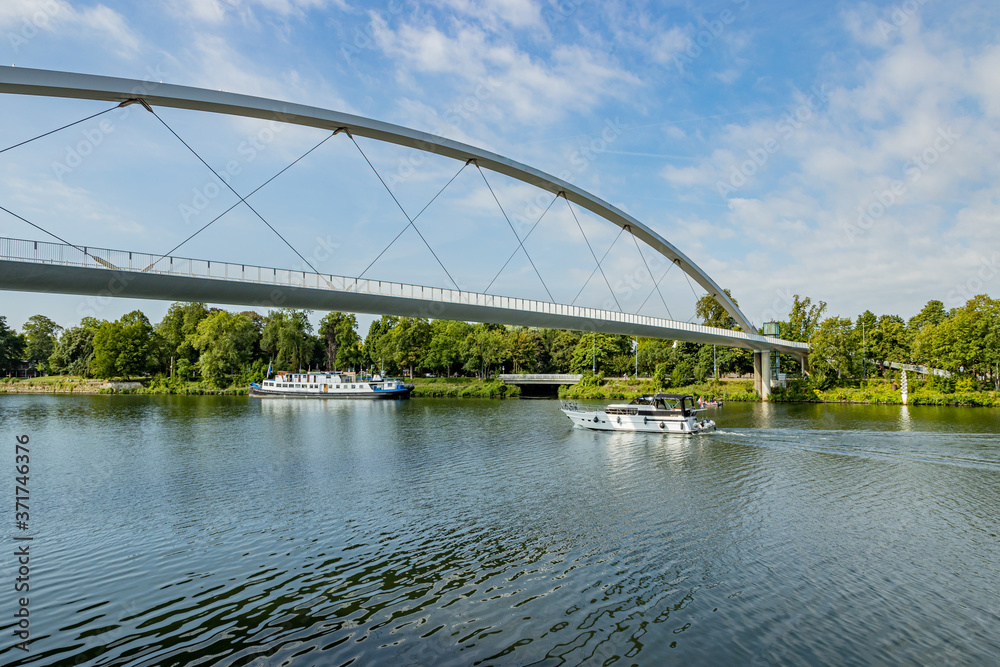 One boat sailing and another anchored on the bank of the Maas river surrounded by green trees and the High Bridge (Hoge Brug), sunny summer day with blue sky in Maastricht, South Limburg, Netherlands