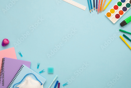 Top view of blue, pink and purple notebooks, pins, eraser, felt tip pens, color pencils, ruler, face mask, marker and watercolor. School stationery set on the blue background. Copy space. 