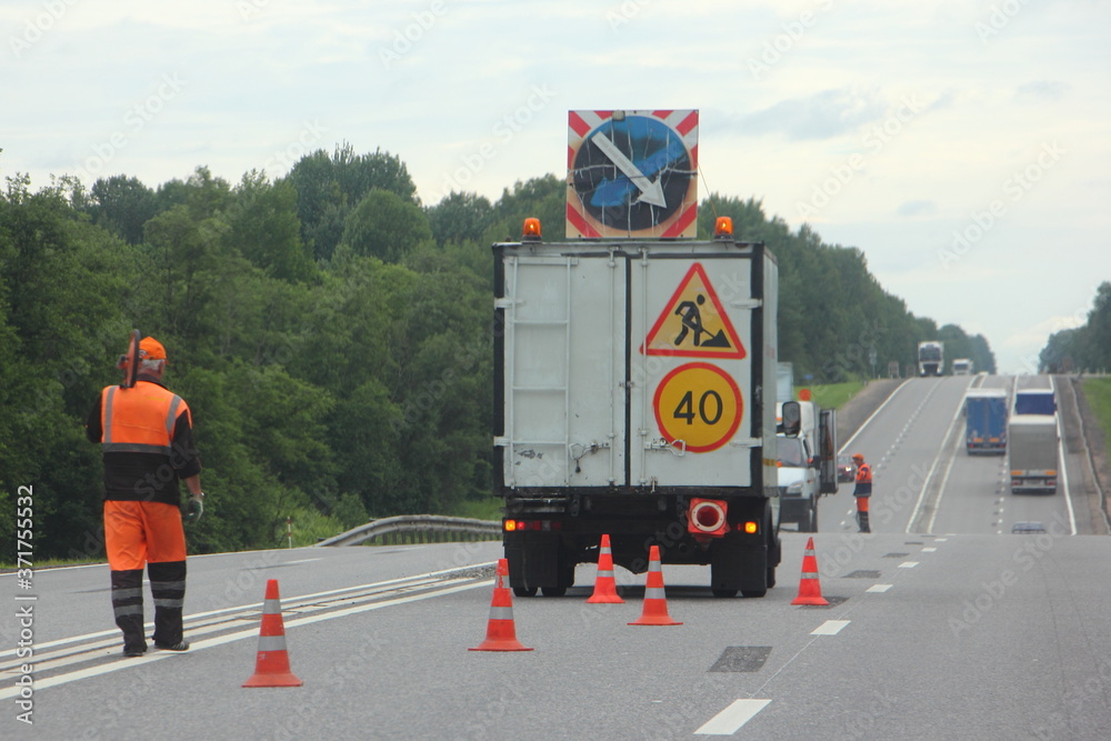 Road repair, horizontal road marking - truck van with workers in orange overalls clothes in the left lane of the road, orange safety cones, direction of detour, sign speed limit 40 km/h and road works