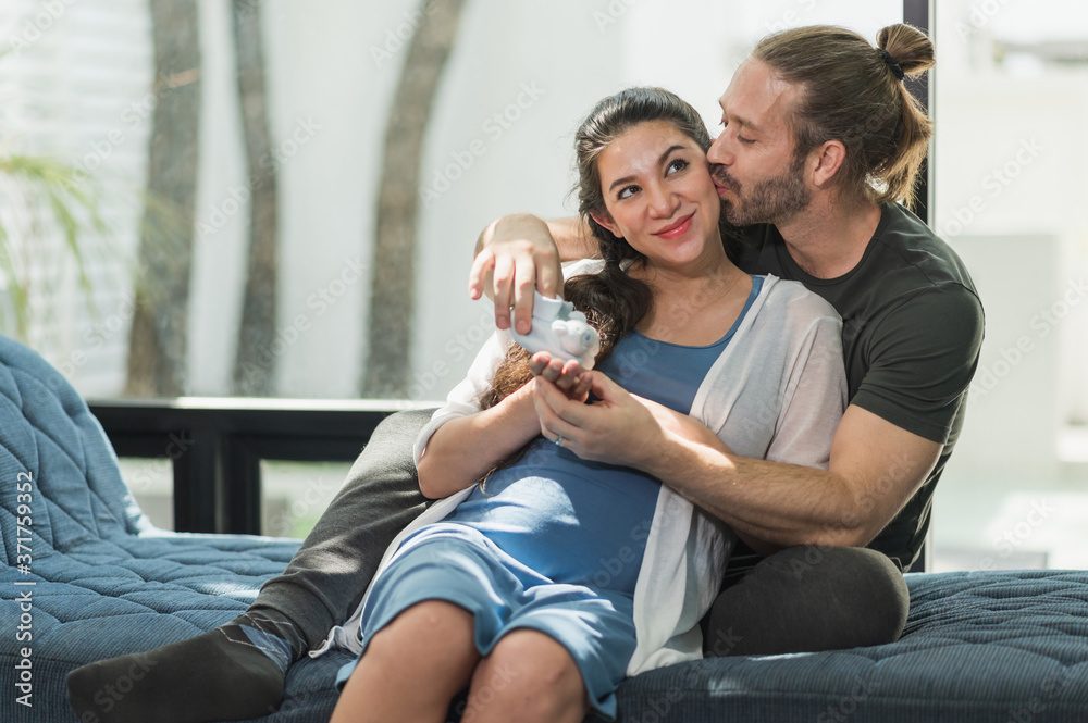 Obraz premium Handsome beard man take care his pregnant wife hugging and holding they shoe baby while sitting on sofa. Happy family concept.