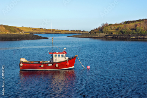 A little red fishing boat lies peacefully at anchor in a sheltered bay on a calm early Spring evening.