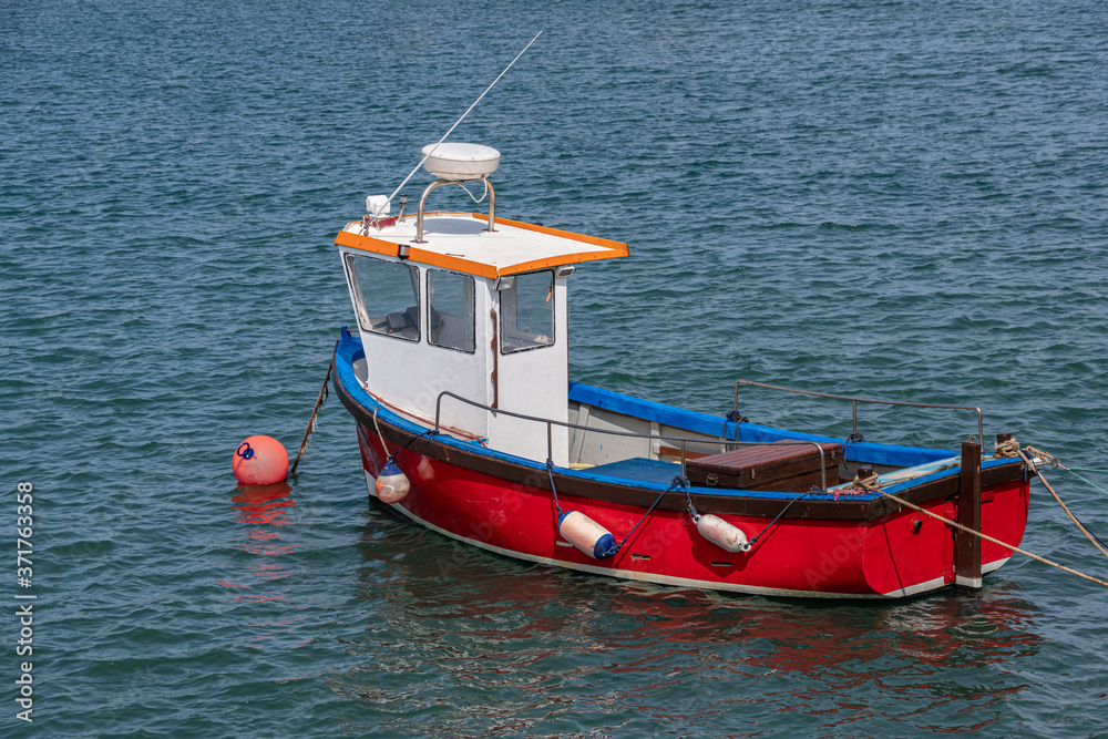 A little red fishing boat lies peacefully at anchor in a sheltered harbor on a warm Summer day.