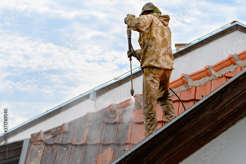 Young professional in yellow uniform diligently performing roof wash with compressed water. After treatment roof tiles cleaned from lichens and dirt look like new ones.