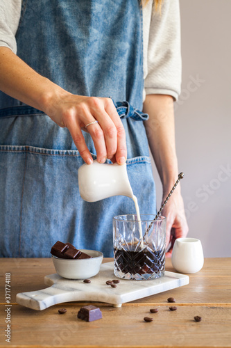 woman pouring milk in coffee glass, coffee beans and chocolate on wooden table