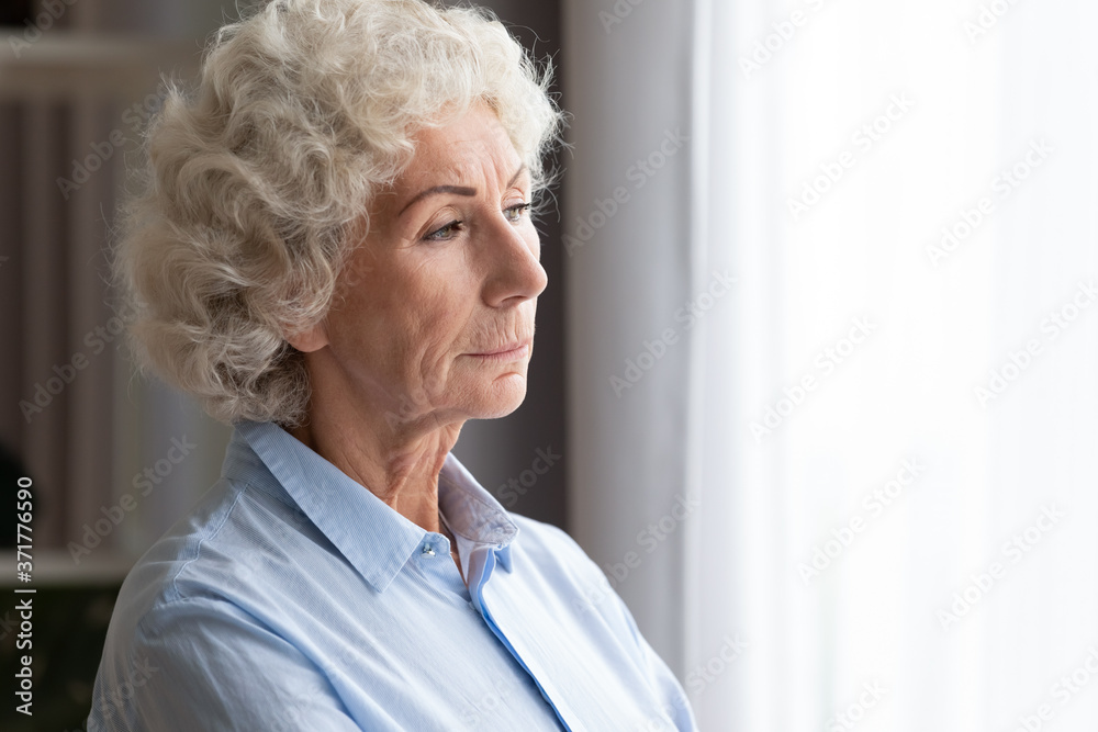 Head shot pensive elderly mature woman standing near window, looking outside contemplating cityscape, thinking of health problems or suffering from mental disease and loneliness at home, copy space. Head shot pensive elderly mature woman standing near window, looking outside contemplating cityscape, thinking of health problems or suffering from mental disease and loneliness at home, copy space.