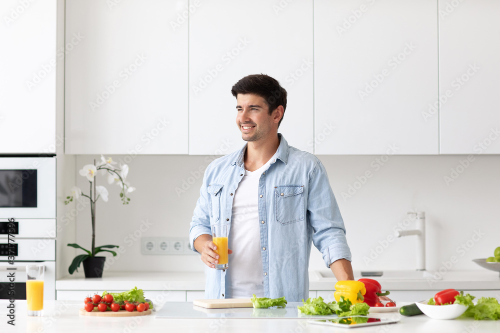 Handsome smiling young man standing in the kitchen home with a glass of orange juice. Vegan food concept.