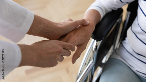 Close up medical social worker holding hand of disabled old retired woman sitting in invalid carriage, giving support or psychological help to handicapped ill patient indoors, rehabilitation concept.
