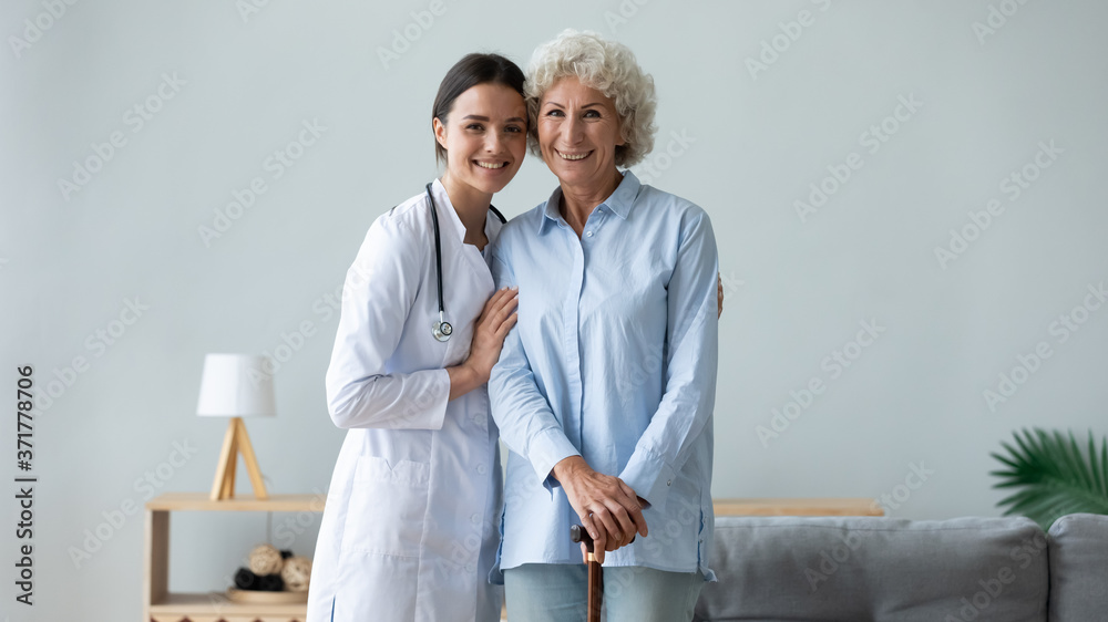 Portrait of smiling young female medical social worker in white coat embracing shoulders of middle aged patient leaning on wooden cane. Happy physiotherapist ang disabled woman looking at camera. Portrait of smiling young female medical social worker in white coat embracing shoulders of middle aged patient leaning on wooden cane. Happy physiotherapist ang disabled woman looking at camera.