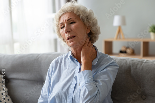 Frustrated middle aged senior woman touching massaging neck, suffering from painful feelings in living room. Unhealthy exhausted mature old grandmother sitting on sofa, feeling muscles tension.