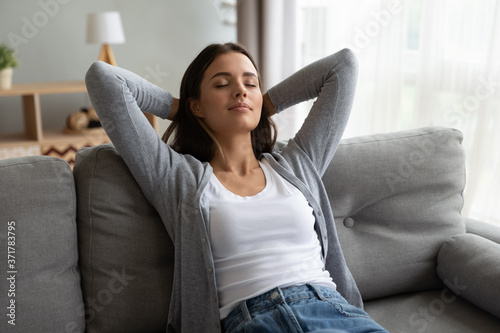 Tableau sur toile Young beautiful peaceful brunette woman leaning on comfortable sofa, stretching back with folded hands behind head, daydreaming with closed eyes, enjoying calm carefree moment alone in living room