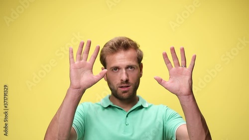 scared young casual man opening mouth in a scary manner, holding hands in the air and surrendering on yellow background