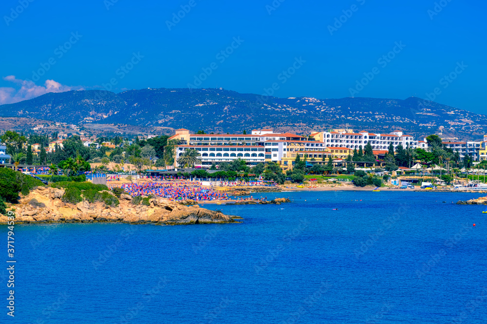 View of a large tourist hotel on the Mediterranean coast of Paphos, Cyprus.  The beach is lined with pink and blue beach umbrellas and is surrounded by cliffs.