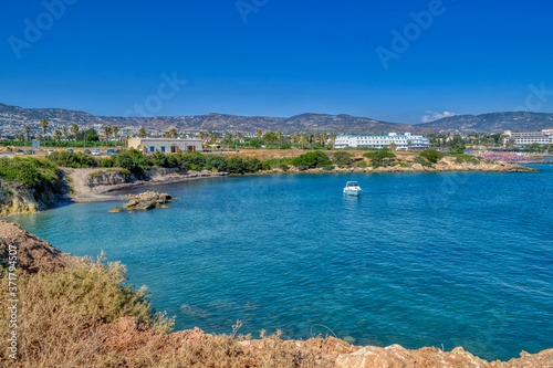 A small lagoon with blue sea water on the coast of Paphos, Cyprus. A small yacht is sailing in the creek. In the distance you can see the hotel with the beach.