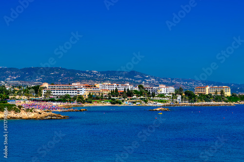 Fototapeta Naklejka Na Ścianę i Meble -  View of a large tourist hotel on the Mediterranean coast of Paphos, Cyprus.  The beach is lined with pink and blue beach umbrellas and is surrounded by cliffs.