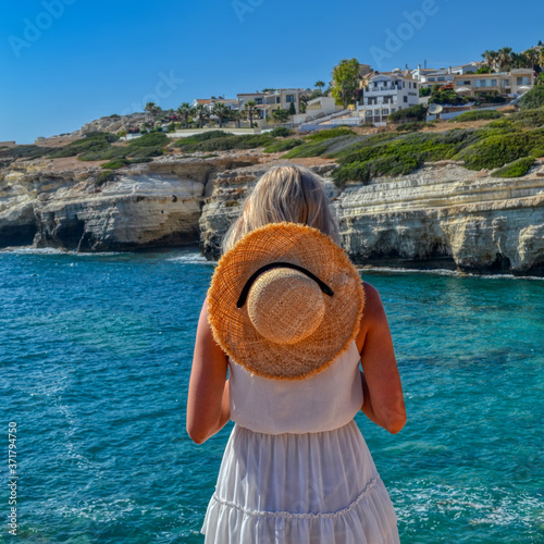 A thin nice girl in a straw hat and a white dress stands on a rock and looks down at the bay with blue sea water. Beautiful woman on a background of Mediterranean cliffs relaxing by the sea. Hot day