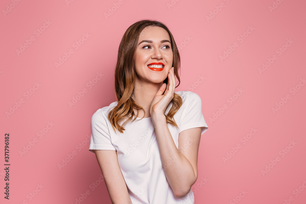 Young caucasian woman with red lipstick and white t-shirt smiling looks at copy space holding hand near cheek isolated on pink background Young caucasian woman with red lipstick and white t-shirt smiling looks at copy space holding hand near cheek isolated on pink background